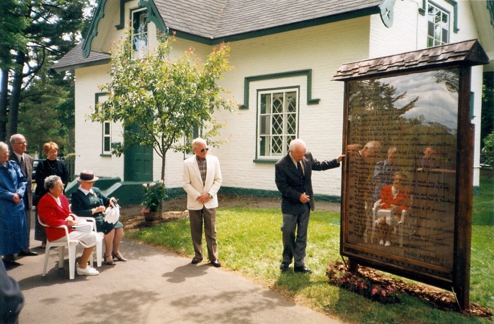 A color picture of the rules sign being presented to a group of people. There is a man presenting the sign, another man wearing a white blazer looking at the sign and women sitting in white chairs wearing nice clothing listening to the presentation. They are people standing in the background in front of the lodge.