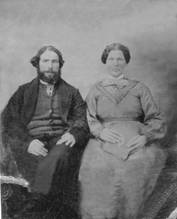 a black and white picture of a couple sitting, wearing period clothing. The woman is on the man right while holding a book.