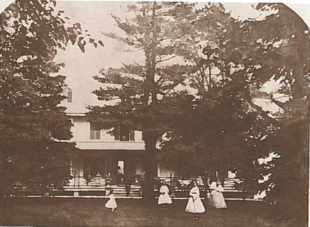 Yellow and orange photo of a house shrouded by conifer trees. There are women dressed in white dresses in front of the house on the grass by a big tree.