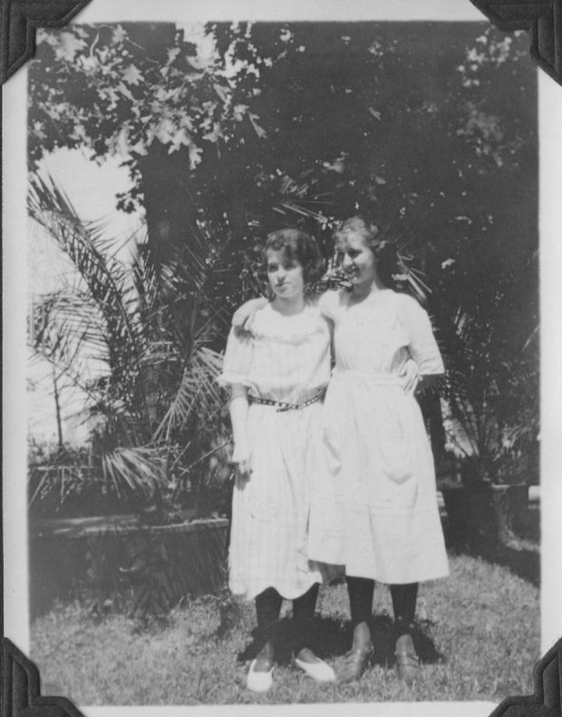 black and white picture two young girls dressed in white in the summer. Surrounded by tropical plants from the greenhouse in the background.