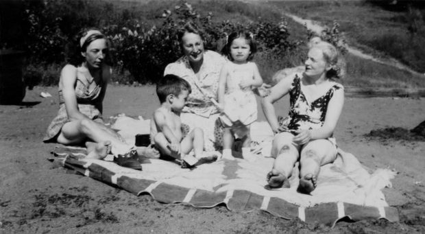 Black and white picture of a group of people sitting on the beach. There are three women dressed in beach attire and a little boy and girl. They are all on a large white sheet.