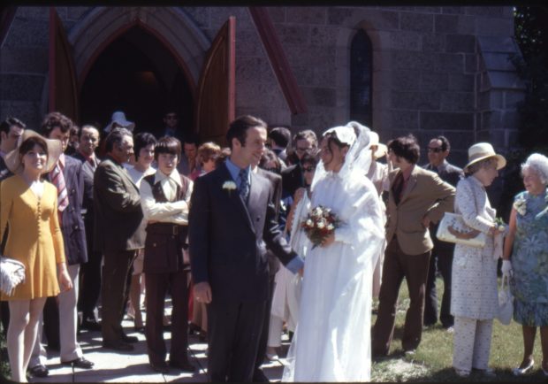 Colour picture of a man and a woman dressed for a wedding. Both coming out of the church they are surrounded by people.
