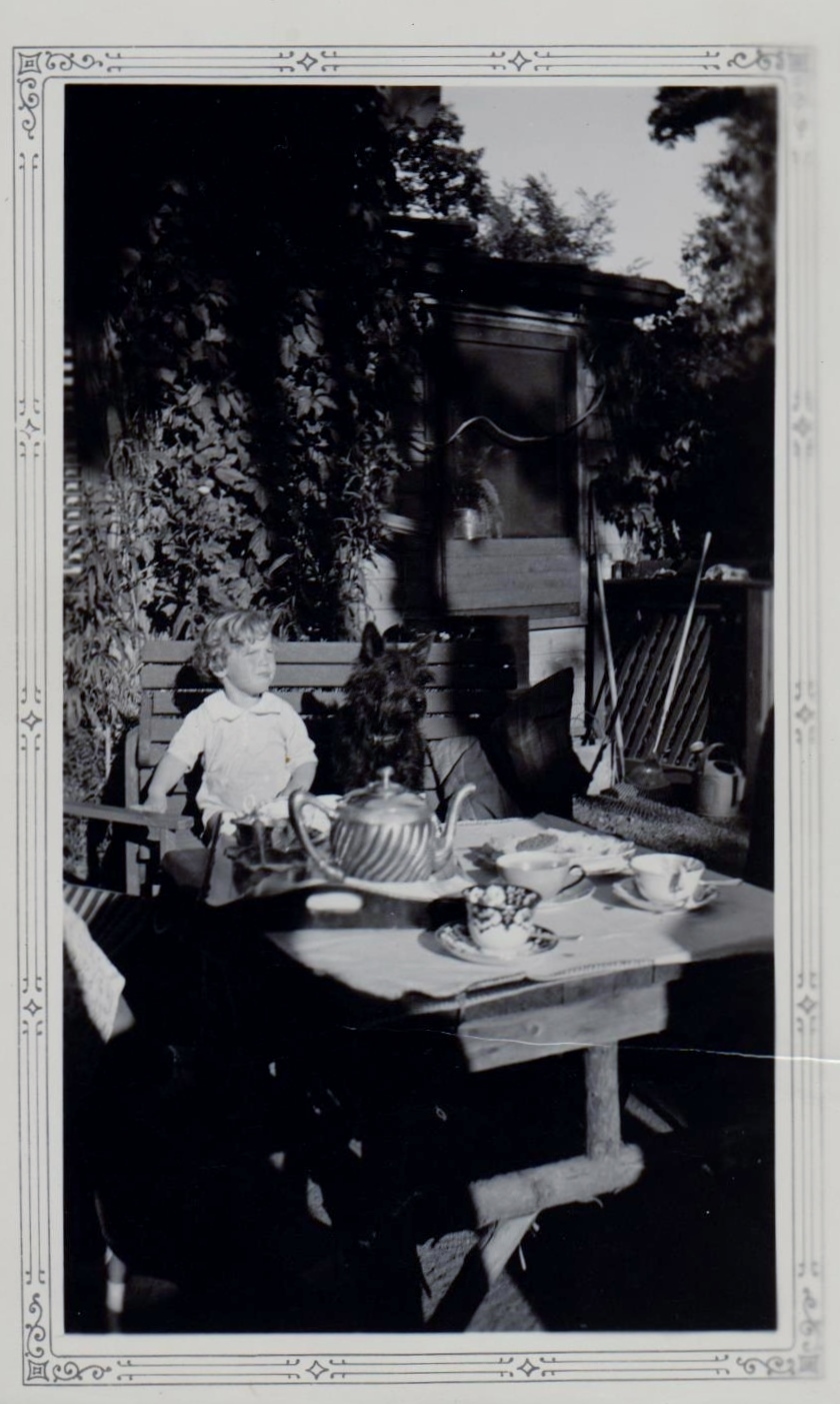 Black and white picture of a little boy and his black dogs sitting on a bench in the garden