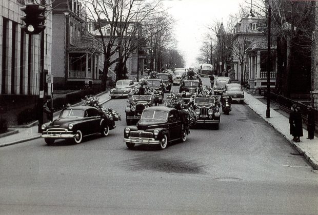 Black and white picture of a funeral procession, many black cars are driving down a road.