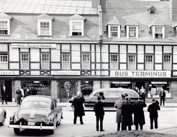 Black and white picture of a funeral procession, many black cars are driving down a road.