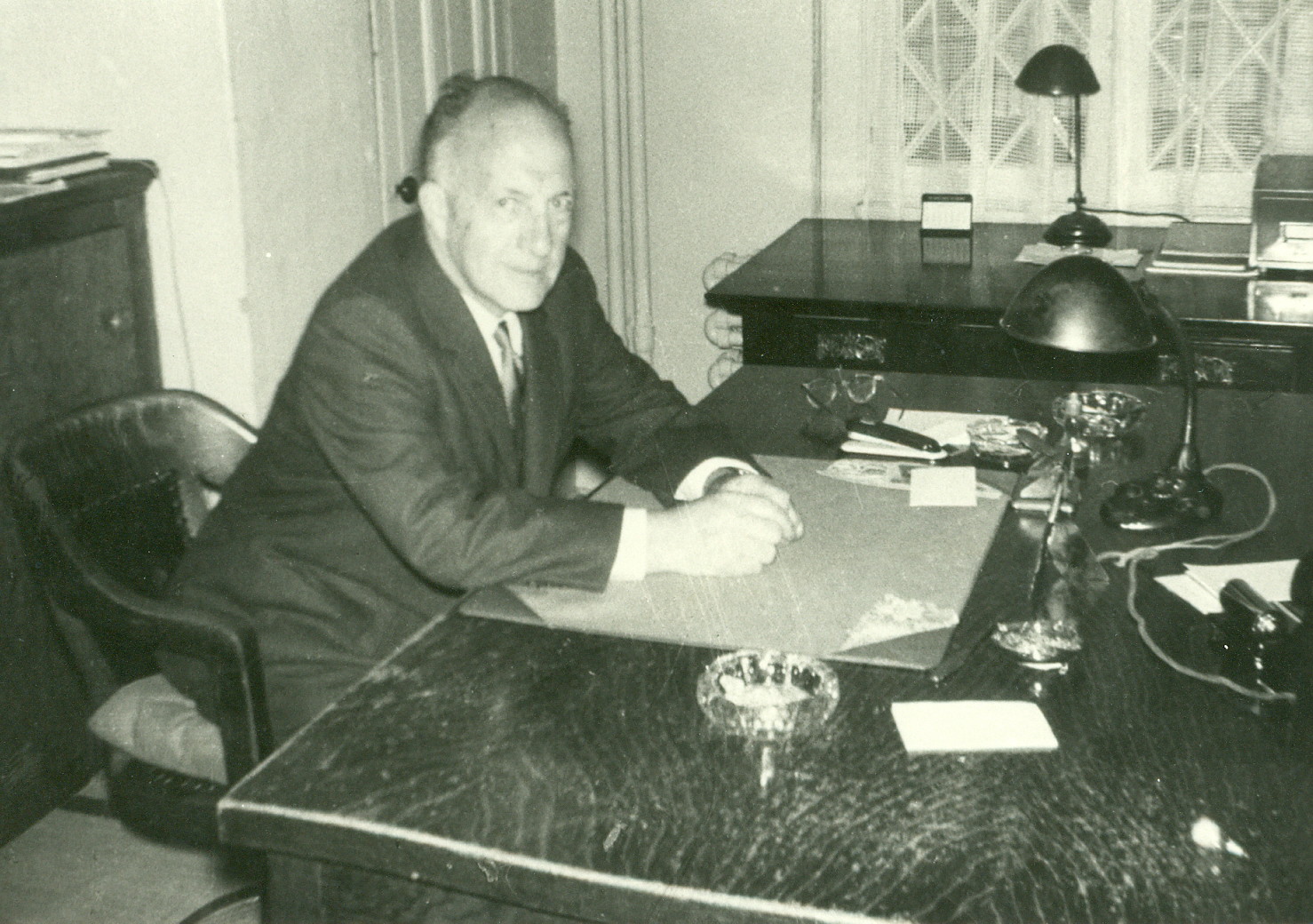 Green and white picture of an old man sitting at a large wooden desk.