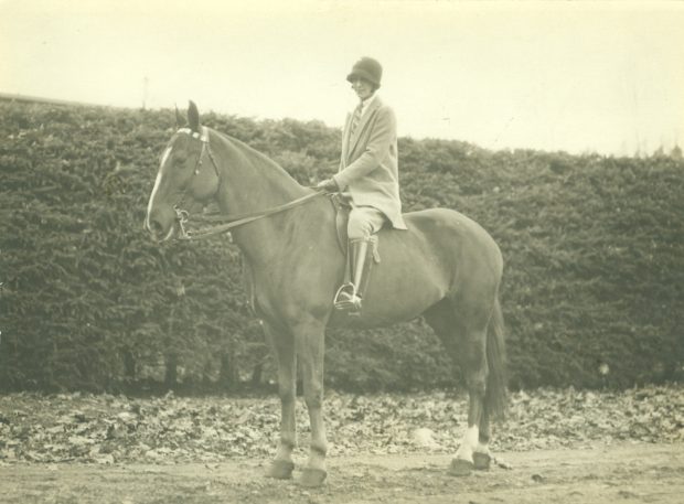 Sepia picture of a woman sitting on a large horse, they are standing in front of a hedge