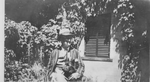 Black and white picture of an old man in a loose fit suit with a pipe in his mouth, with a black Scottie dog on his lap. They are sitting on a chair in front of the house, and house vines surround them.