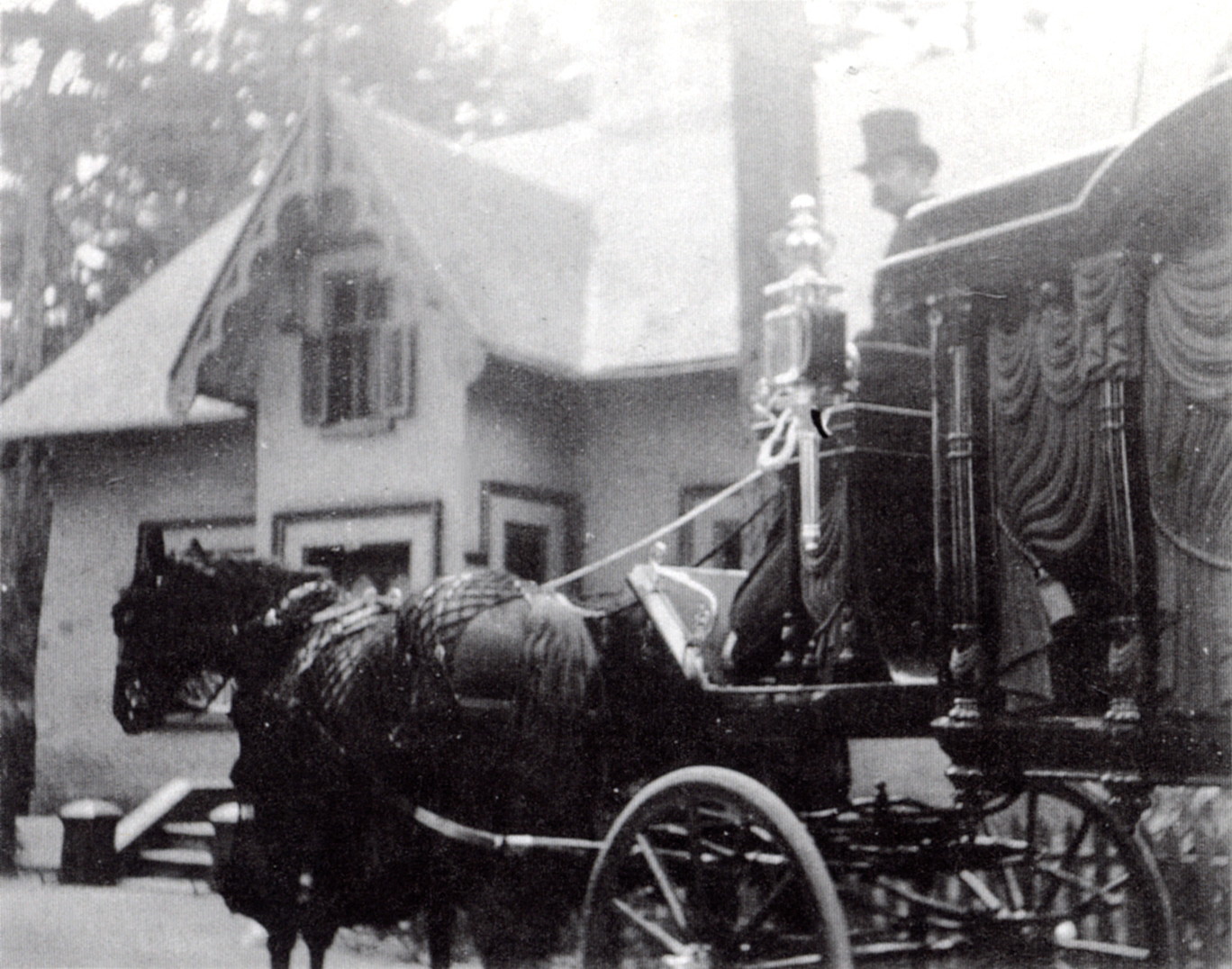 Black and white picture of a funeral procession, with a horse-driven hearse and a man wearing a top hat