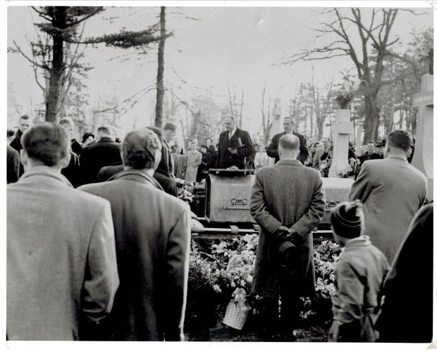 Black and white picture of a burial service. Men are standing with their head bowed around a casket lowered into the ground with a curious little boy in the background. Flowers and granite headstone surround the casket.
