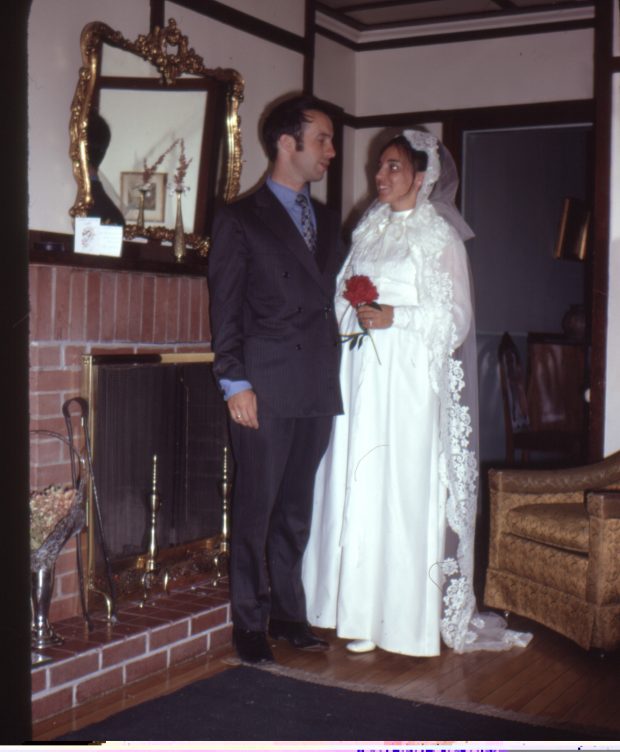 Colour picture of a man and a woman dressed for a wedding. They are standing in front of a fireplace.