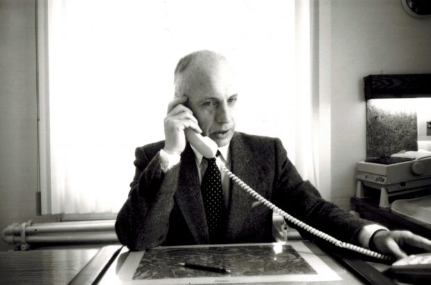 Black and white picture of a man dressed in a suit holding a phone to his ear in a office