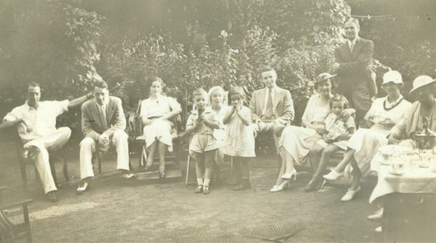 A sepia picture of men, women and children sitting in a semi-circle wearing lavish clothing at a Treggett family tea party surrounded by a lush scenery of green vines
