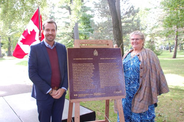 Man and woman standing on either side of a commemorative plaque for the National Historic site of Canada with a Canadian flag in the background