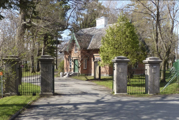 Coloured picture of a brick house surrounded by trees and a metal fence with an opening with four large cement post for a beautiful driveway.