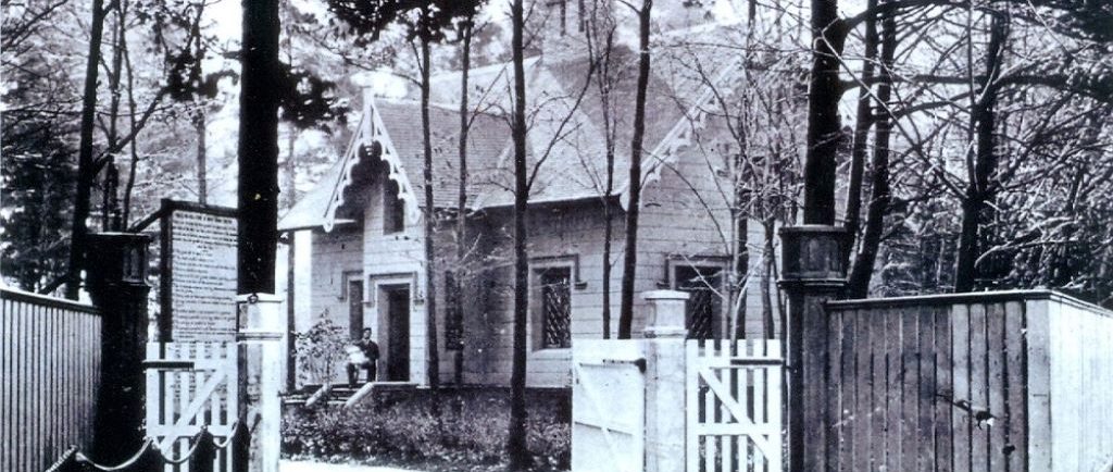 Black and white picture of a house in the woods. A man is sitting on the front step holding a white-clothed baby. There is also a large wood fence in front of the house.