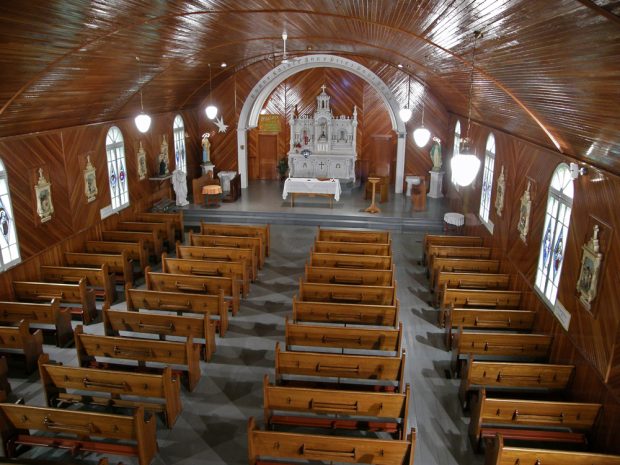 Color photograph, wide shot of inside of church, both ceiling and walls are covered with varnished wood, with stained glass windows, all religious furnishings are in place.