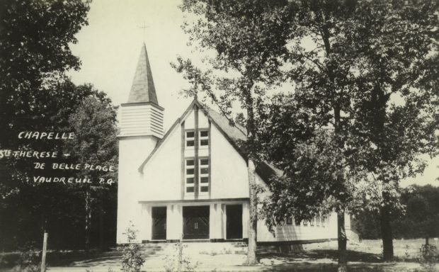 Old black and white photograph taken in summer, close-up of a white wood church facade, the steeple is placed at the top of a tower on the church’s left side.