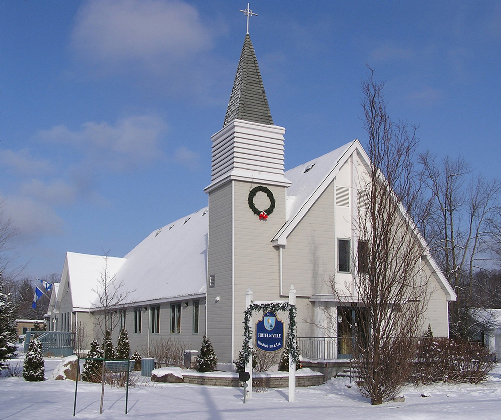 Color photograph taken in winter, close-up of a white wood church facade, the steeple is placed at the top of a tower on the church’s left side, in front, a sign reads: City Hall.