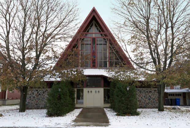 Color photograph taken in winter, close-up of church facade on which the lower part of the walls is covered in stones, the gabled roof contains stained glass windows and there are trees on either side.