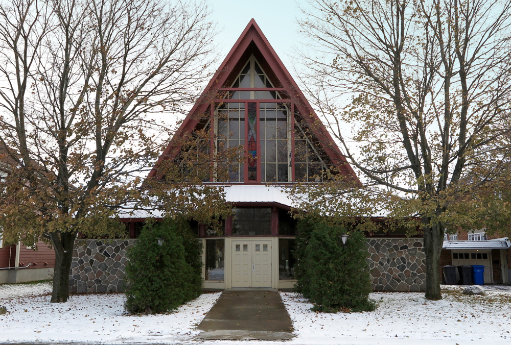 Color photograph taken in winter, close-up of church facade on which the lower part of the walls is covered in stones, the gabled roof contains stained glass windows and there are trees on either side.