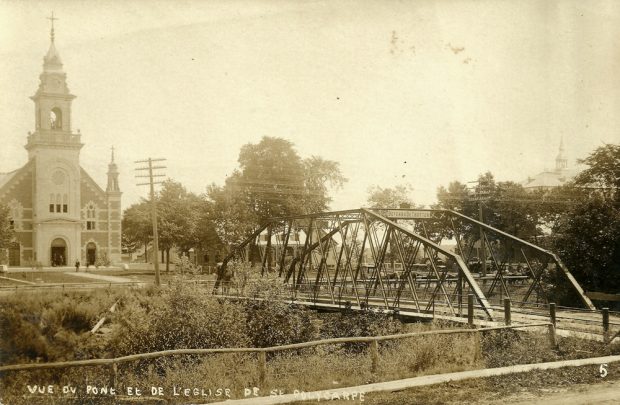 Old black and white photograph, long shot of a church facade facing a road and metal bridge.