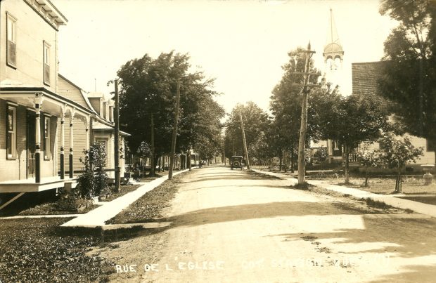 Black and white photograph, long shot of a village street, on each side are houses and a sidewalk, in the background a church steeple is visible above the trees.
