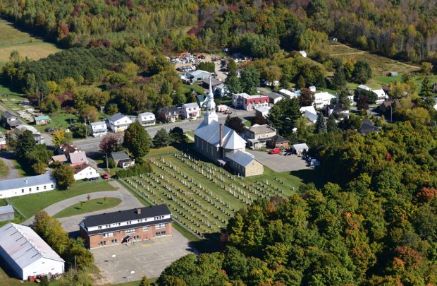 Color photograph, aerial view of a village, in the center, a church and cemetery surrounded by houses and buildings, enveloped by a forest.