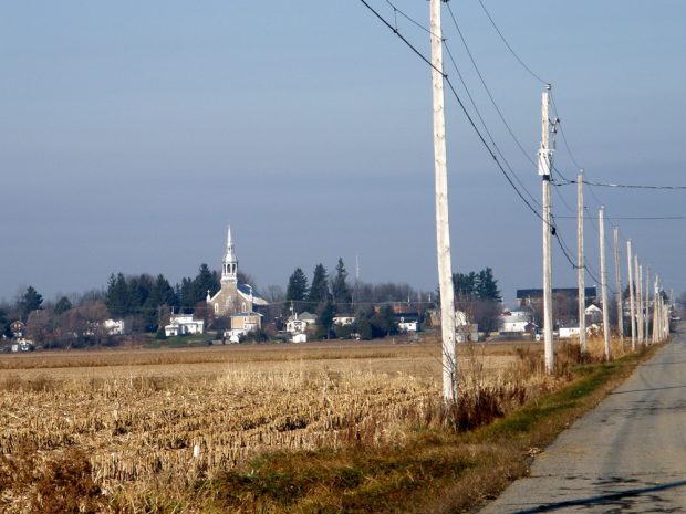 Color photograph, long shot of a church in the center of a village, in the foreground, a field and electric poles bordering a paved road.