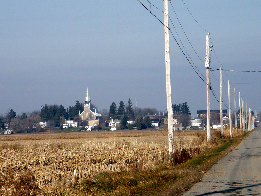 Photographie couleur, plan éloigné, d’une église au centre d’un village, en avant-plan un champ et des poteaux électriques longeant une route asphaltée.