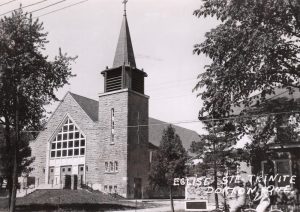 Old black and white photograph of a stone church facade and presbytery, in the foreground on the right, two young girls are riding bicycles.