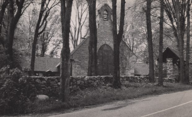 Old black and white photograph, stone church facade surrounded by trees, in the foreground, a low stone wall borders a paved road.