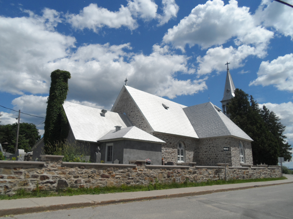 Color photograph, side view of a stone church with metal multi-sloped roof and a steeple, surrounded by a low stone wall bordering a sidewalk and paved road.