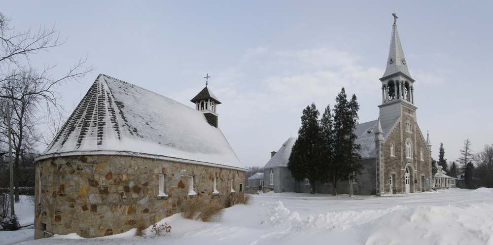 Color photograph, long shot taken in winter, in the foreground, back view of a small stone chapel with tiny steeple topped with a cross, in the background, side-view of a stone church with a steeple, a low stone wall and the facade of a stone presbytery.