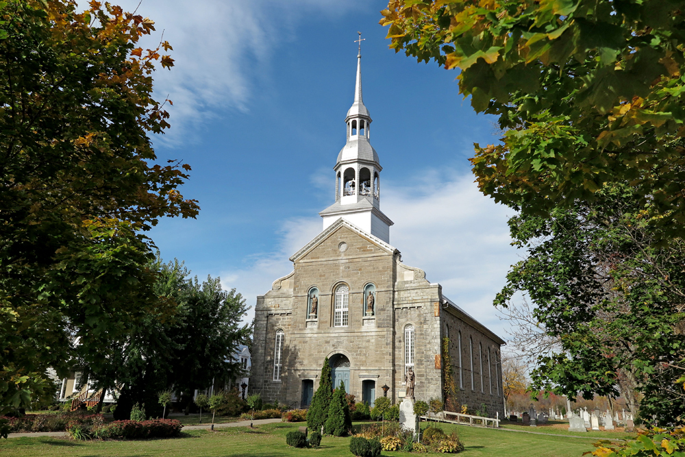 Photographie couleur, plan éloigné, façade d’une grande église en pierres taillées  surmontée d’un clocher en bois et en tôle, entourée d’arbres. En arrière plan, un presbytère à gauche et un cimetière à droite. 