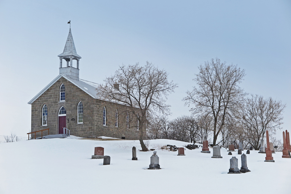 Color photograph, wide shot taken in winter, frontage of a stone country church, to the right, its cemetery and trees.