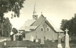 Photographie ancienne en noir et blanc, plan éloigné, vue d’un cimetière entouré d’un muret et d’arbres, situé à l’arrière d’une église en crépi dont la toiture est à plusieurs versants avec un clocher. 