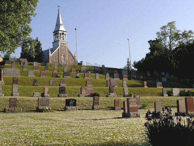 Color photograph, long shot, in the foreground, a cemetery built on multiple levels of a cliff, in the background, a stone church façade and steeple.