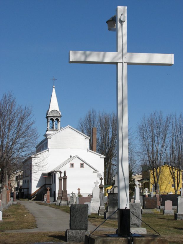 Color photograph, back view of a church, in the foreground, a cemetery and a big white cross.