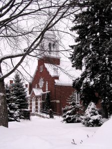 Color photograph taken in winter, side view of a red brick church with a wood steeple covered with sheet metal, in the foreground, a path leading to the main entrance and snow-covered trees.