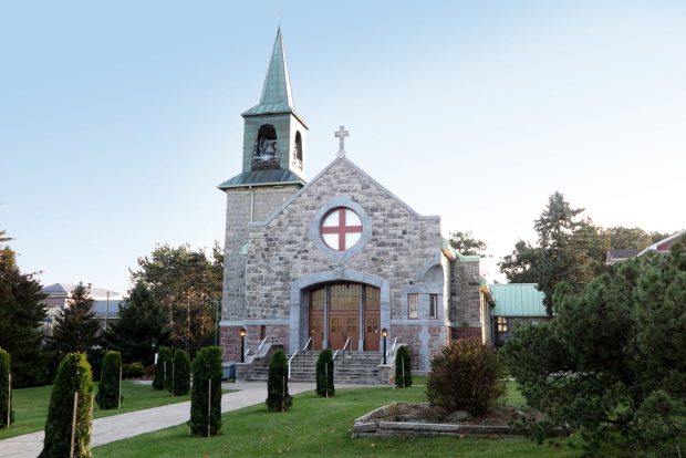 Color photograph, long shot, stone church façade with cross on top, to the left, the stone steeple tower and oxidized copper roof, and landscaping in the foreground.