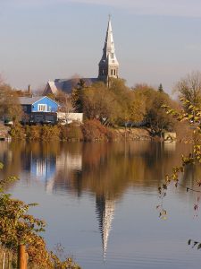 Color photograph, long shot of a church steeple, a house and trees on the waterfront and their reflection on the water.