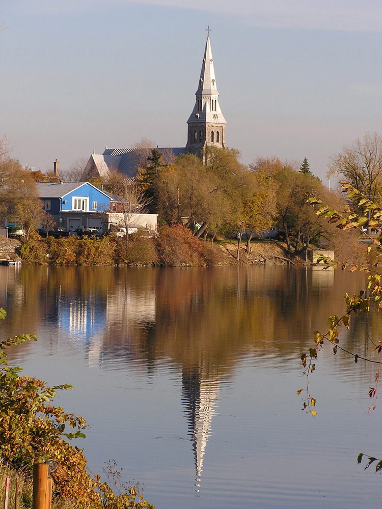 Color photograph, long shot of a church steeple, a house and trees on the waterfront and their reflection on the water.