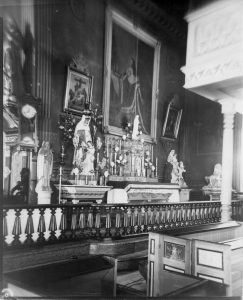 Old black and white photograph, close-up of church interior, richly decorated chapel with liturgical furnishings, statues and paintings, in the foreground, a varnished wood railing and pews.