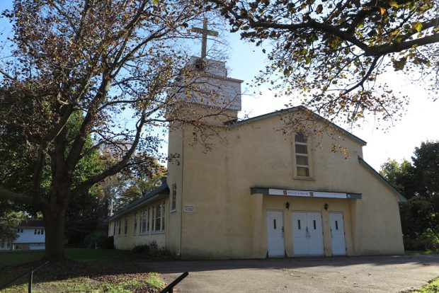 Color photograph, close-up of a yellow-painted cement church façade, to the left, a square steeple topped with a white wooden cross.