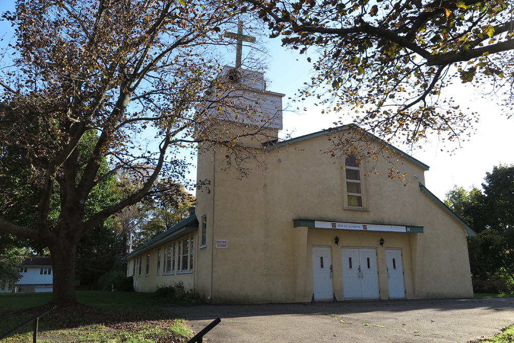 Color photograph, close-up of a yellow-painted cement church façade, to the left, a square steeple topped with a white wooden cross.