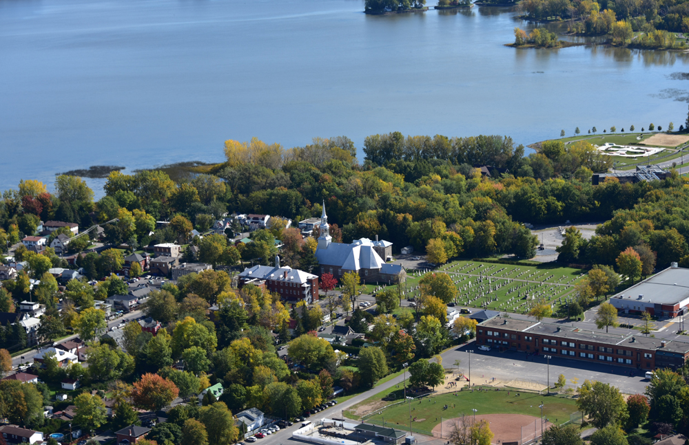 Color photograph, aerial view of a city on a lakeside, in the center, a church and cemetery, on the left, houses and trees, on the right, a park and a school.