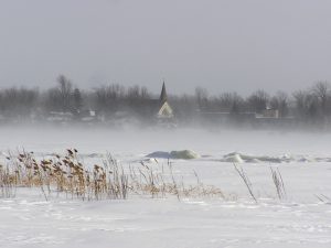 Color photograph taken in winter, long shot of a village on a frozen waterfront, in the center, a church steeple is visible through the low drifting snow.