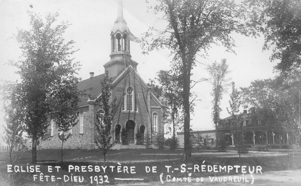 Old black and white photograph, facade of a stone church and presbytery surrounded by trees, the church is decorated with ribbons attached to the steeple and flowing down to the church steps.