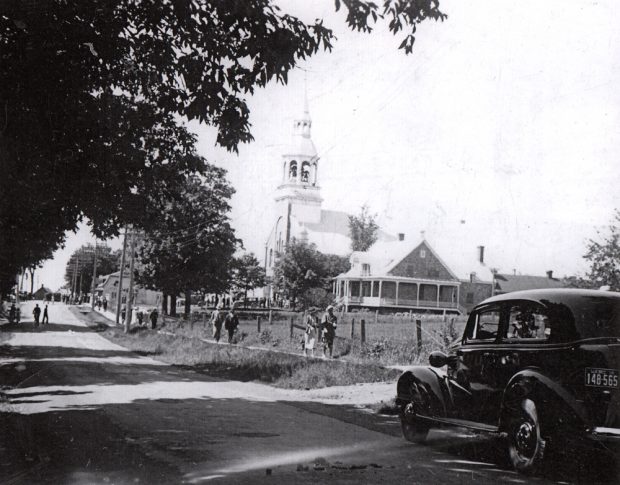 Old black and white photograph, in the foreground, a road, car and people walking on the sidewalk, in the background, a church steeple and presbytery.